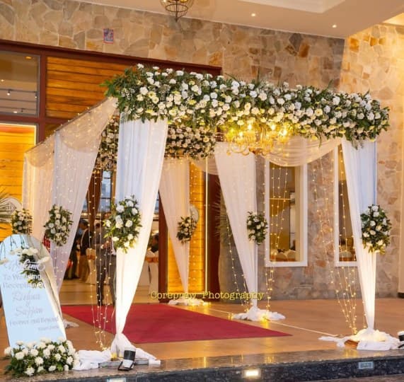 Venue entrance with a white floral canopy, ivory drapes, string lights and a welcome sign