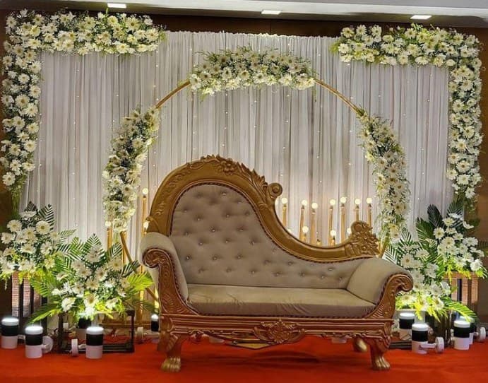Gold ring arch covered in white gerberas behind a carved gold chaise, with candles and greenery