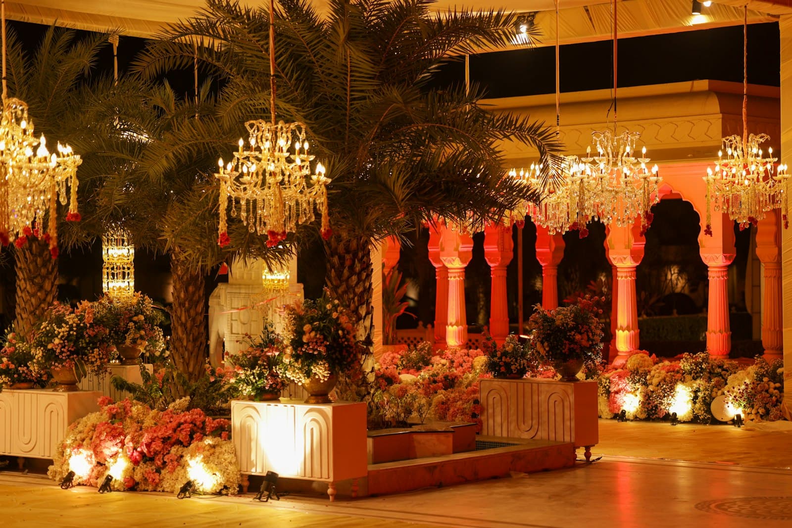 Evening reception venue lit by chandeliers hanging from palm trees, with floral pillars