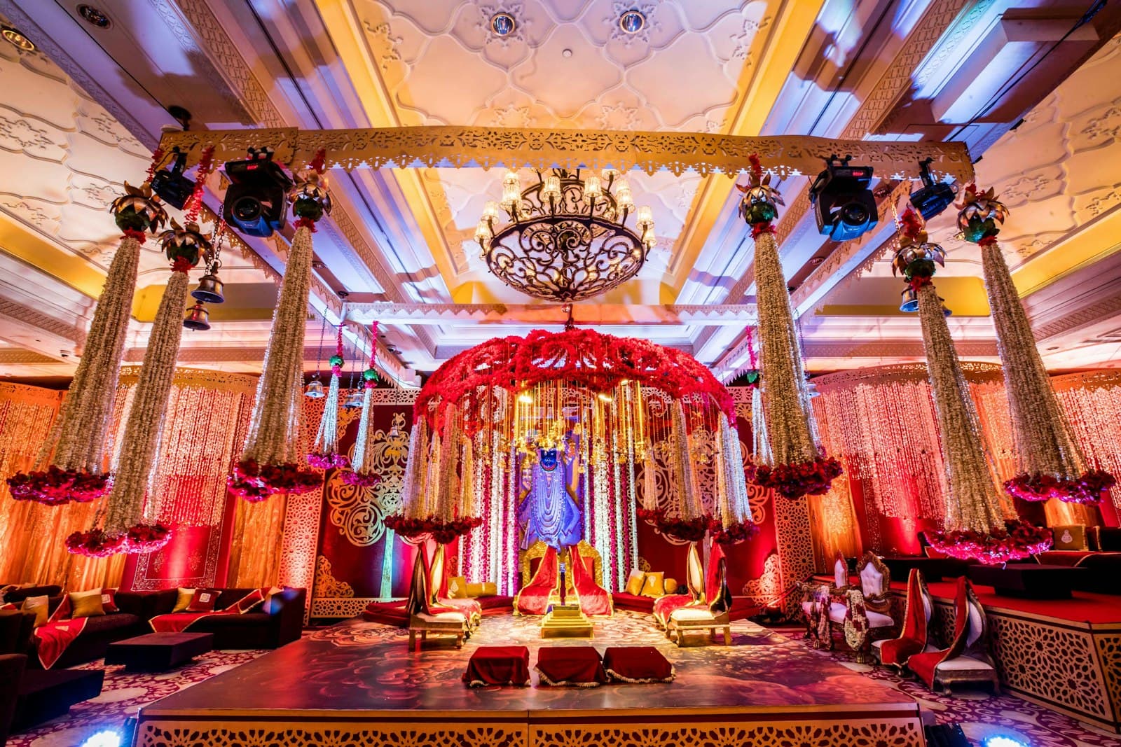 Grand indoor mandap with tall floral pillars, red drapes and a crystal chandelier
