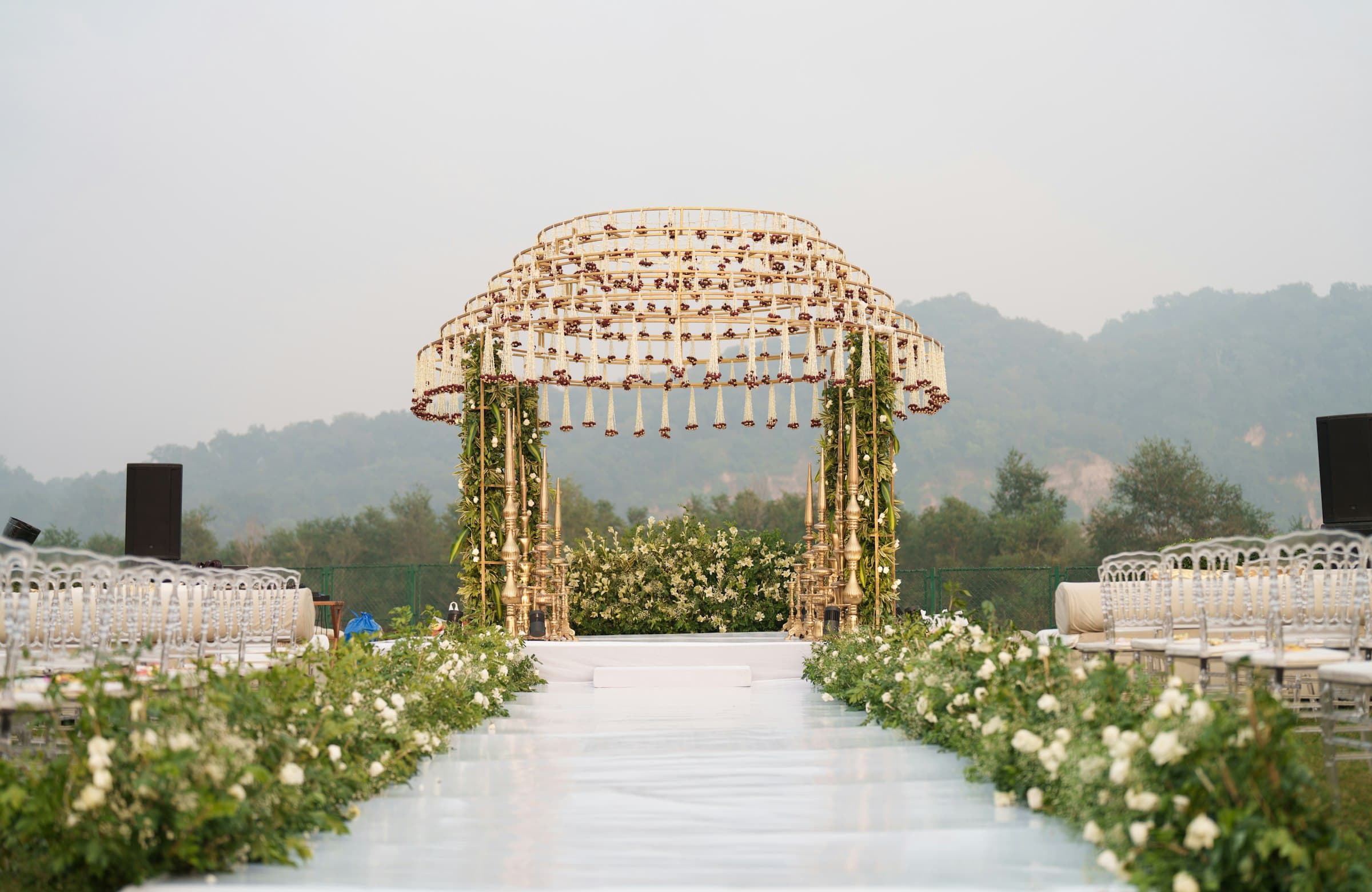 Open-air wedding mandap with a gold canopy of hanging jasmine strands, a white aisle lined with white roses and hills behind